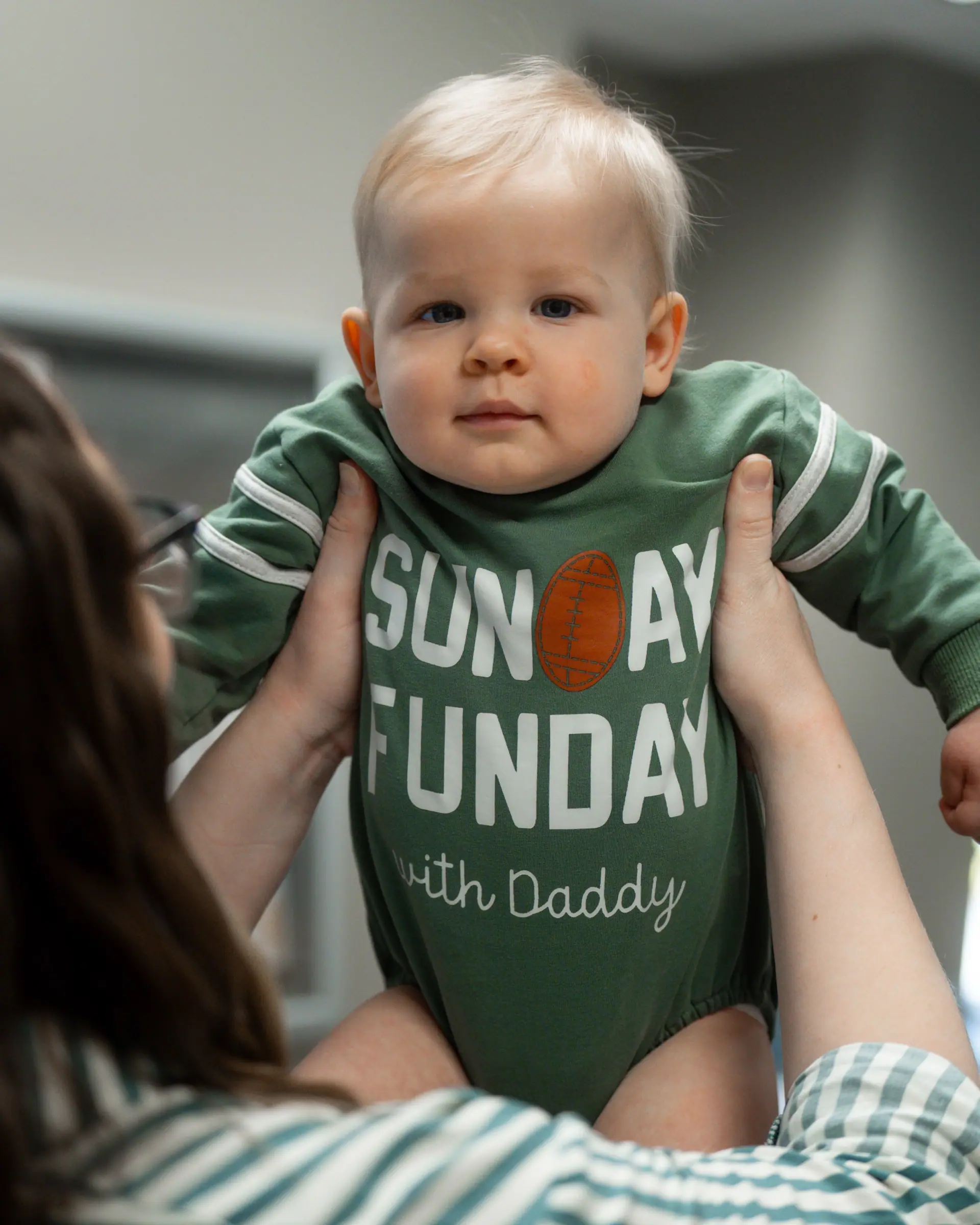 A baby in a Sunday Funday shirt held up by his dad